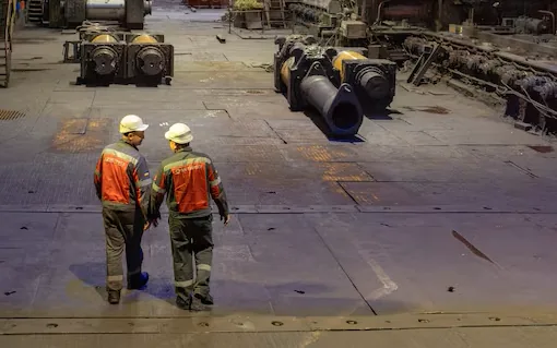 Two workers in high-visibility clothing crossing a steel mill floor